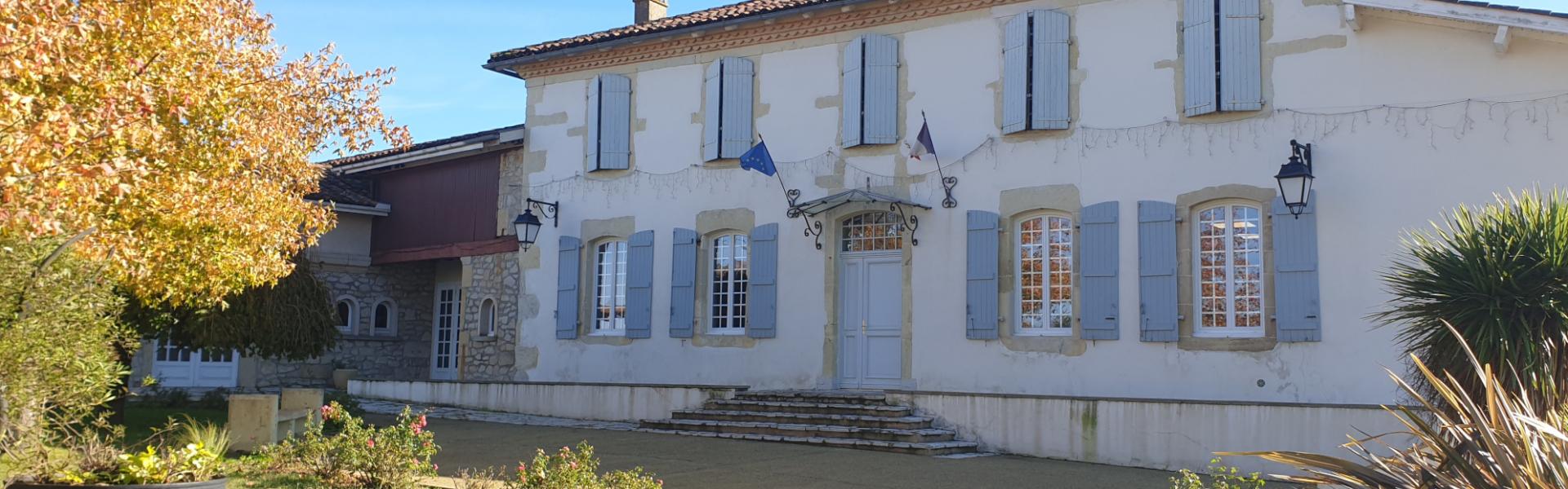 Photo de la Mairie de Laurède. C’est un bâtiment de style traditionnel, pavoisé avec les drapeaux français et européen fixés en façade. Les volets bleu clair et les pierres apparentes sont typique de l'architecture locale. Le jardin est fleuri, des arbustes entoure le bâtiment.